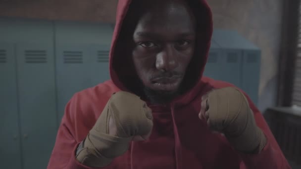 Крупным планом портрет лица и рук молодого афроамериканского боксера в красной толстовке, с обёрнутыми руками на сжатых кулаках, стоящего на страже в раздевалке и смотрящего в камеру - Кадры, видео