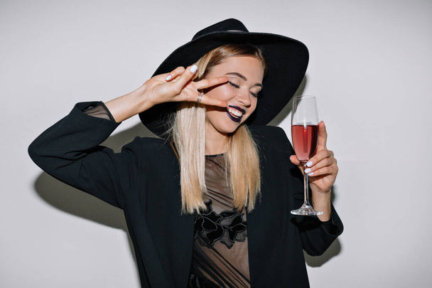 Funny excited girl with blond hair wearing black holiday outfit dancing and showing peace sign with a glass of wine over isolated background . High quality photo - Photo, Image