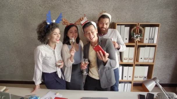 Monietninen ryhmä nuoria naisia ja miehiä, jotka ottavat selfien älypuhelimella ja hymyilevät iloisesti viettäessään joulua virassa - Materiaali, video