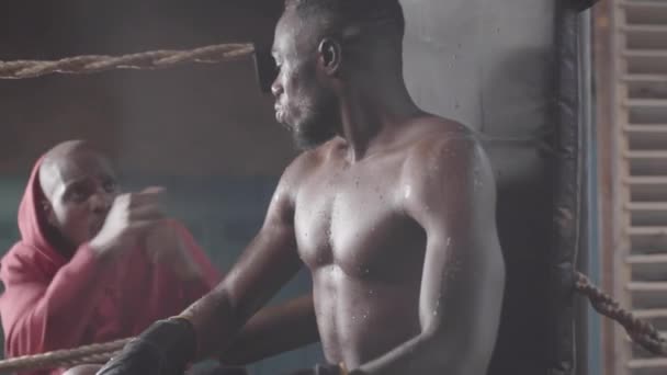 Taille-up shot van bezwete gespierde Afro-Amerikaanse bokser zittend in ring hoek tussen match rondes, drinkwater en luisteren naar opgewonden mannelijke coach, die wijst en geeft instructies - Video