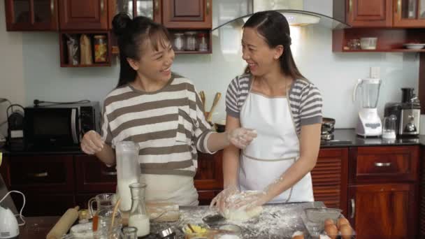 Houkutteleva aasialainen nainen vaivaa evästetaikinaa, kun hänen kaunis tyttärensä lisää jauhoja, pölyttää kätensä ja hymyilee onnellisesti. - Materiaali, video