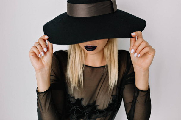 Indoor close-up portret van charmante jonge vrouw met hoed en zwarte blouse die lacht en haar hoed vasthoudt. Hoge kwaliteit foto - Foto, afbeelding