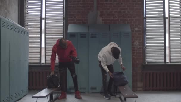 Komplette Aufnahme von zwei freundlichen jungen afroamerikanischen Sportlern, die sich in der Umkleidekabine des Fitnessstudios unterhalten, Boxhandschuhe aus der Umhängetasche ziehen und sich gegenseitig zeigen - Filmmaterial, Video