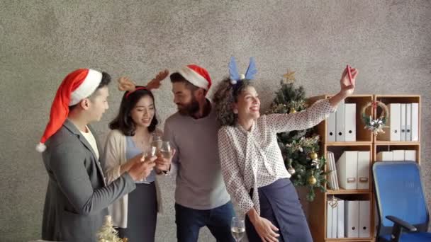 Gruppo multietnico di giovani allegri impiegati maschi e femmine che festeggiano il Natale al lavoro, bevono bicchieri di champagne e scattano selfie sullo smartphone - Filmati, video