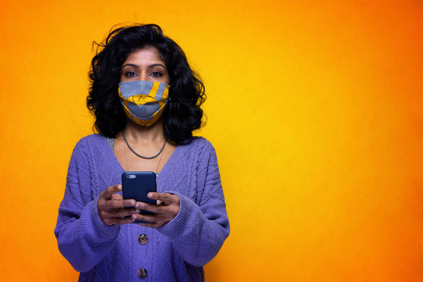 Csinos indiai lány SMS-ezik és játszik a telefonján, kovid 19 füstmaszk - Gyönyörű Sti Lanka fiatal nő használja a telefont, hogy csevegés és vásárlás online visel koronavírus pandémiás védelem - Fotó, kép