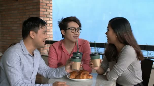 Mediana toma de tres compañeros de trabajo sentados en el café con tazas de papel de café en sus manos y hablando - Metraje, vídeo