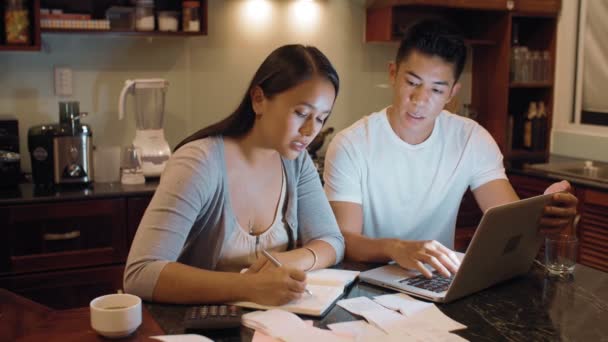 Joven pareja asiática casada administrando las finanzas en casa, sentado en la mesa de la cocina, calculando los gastos en el ordenador portátil y anotando los números al libro del presupuesto   - Metraje, vídeo