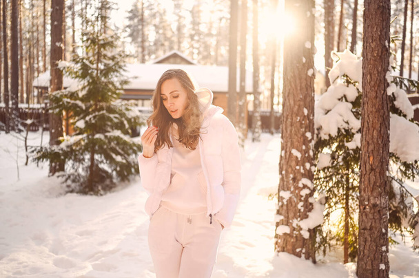 Winter sunny cold portrait in snowfall of beautiful young woman in a white warm outwear down jacket in backyard of country wooden house on the background of green spruce pine trees covered with snow. - Photo, Image