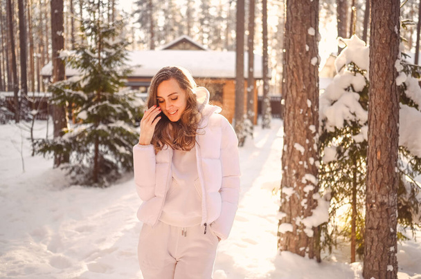 Winter zonnig koud portret in de sneeuw van mooie jonge vrouw in een witte warme outwear donsjas in de achtertuin van het land houten huis op de achtergrond van groene sparren pijnbomen bedekt met sneeuw. - Foto, afbeelding