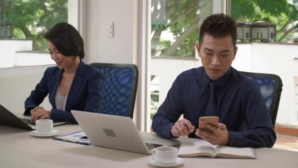 Plan moyen de jeune homme d'affaires asiatique au premier plan travaillant avec un ordinateur portable, prenant le téléphone, faisant appel aux affaires et écrivant quelques informations, et jeune femme d'affaires près de lui en utilisant un ordinateur portable - Séquence, vidéo