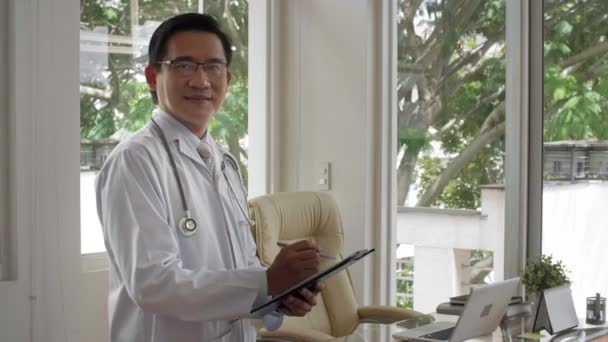 Itsevarma keski-ikäinen mies lääkäri seisoo klinikan konsultointihuoneessa, pitäen leikepöytää ja hymyillen kameralle onnellisena - Materiaali, video