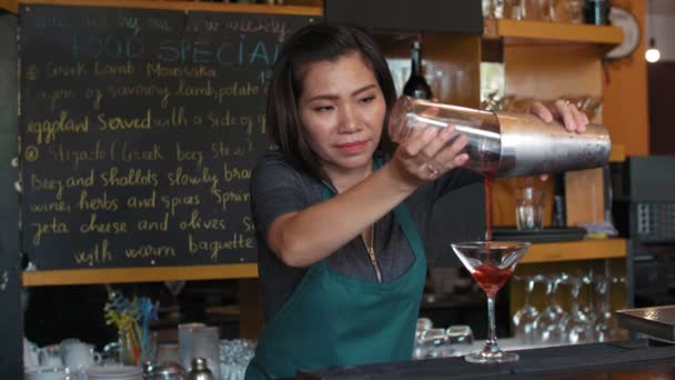 Профессиональная азиатская барменша наливает алкогольный коктейль из шейкера в стекло и украшает его лимоном, стоя у барной стойки - Кадры, видео