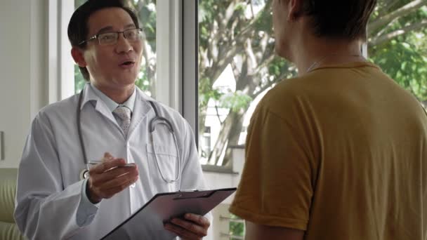Profesionální mužský asijský lékař pozorně naslouchá nepoznatelnému mužskému pacientovi, zapisuje si poznámky do schránky a usmívá se - Záběry, video