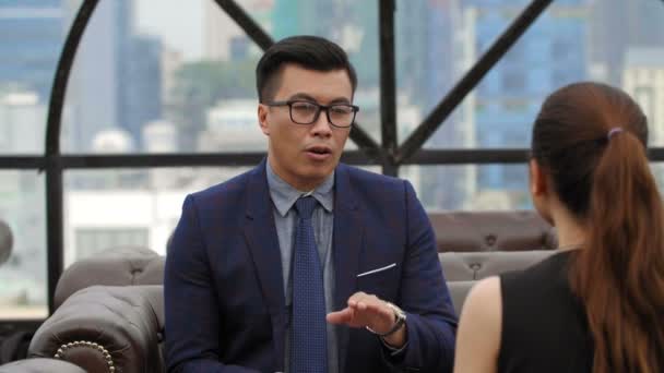 Aussperrung einer asiatischen Geschäftsfrau und eines Geschäftsmannes, die auf einem Sofa vor einander sitzen und miteinander reden - Filmmaterial, Video