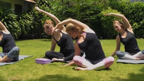 Tracking shot van jonge vrouwen zitten op fitnessmatten en het doen van side bend oefening tijdens het beoefenen van yoga met leraar op groen gras outdoor - Video