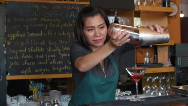 Cantinera asiática profesional que vierte cóctel alcohólico de coctelera en vaso y lo adorna con limón mientras está de pie en el mostrador del bar - Imágenes, Vídeo