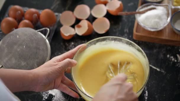 Close-up widok kobiet ręce mieszanie ciasta ciastko z trzepaczką na niechlujny stół kuchenny z skorupkami jaj i składników do pieczenia - Materiał filmowy, wideo
