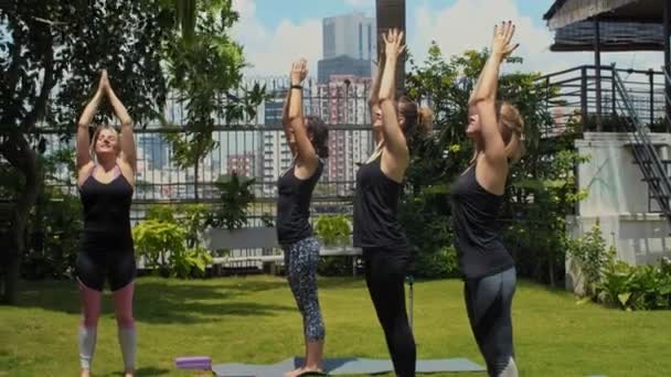 Bir grup genç kadın spor paspasları üzerinde duruyor, kollarını başlarının üzerine kaldırıyor ve sonra dışarıda kadın eğitmenle yoga yaparken öne doğru eğiliyorlar. - Video, Çekim