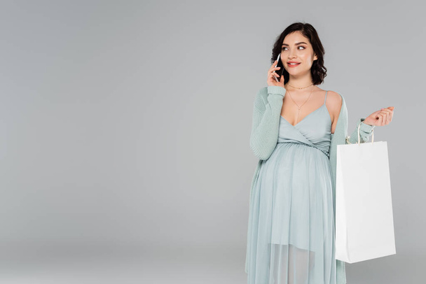 Fröhliche Schwangere spricht auf Smartphone und hält weiße Einkaufstasche isoliert auf grau  - Foto, Bild