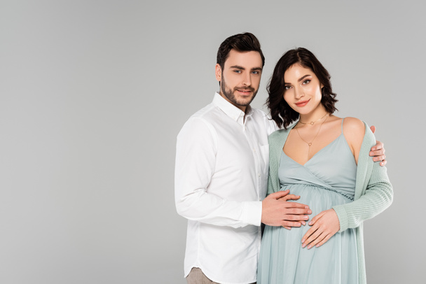 Smiling man in white shirt embracing pregnant wife isolated on grey  - Φωτογραφία, εικόνα