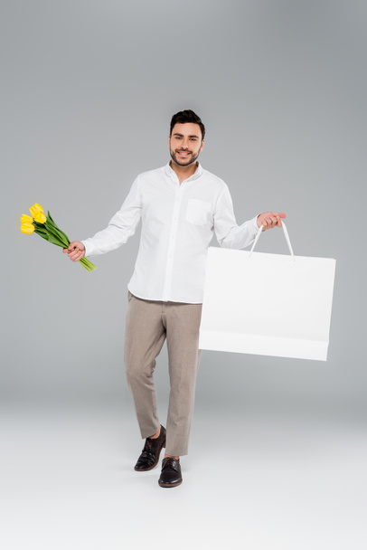 Fröhlicher Mann mit Tulpen und Einkaufstasche blickt in die Kamera auf grauem Hintergrund - Foto, Bild