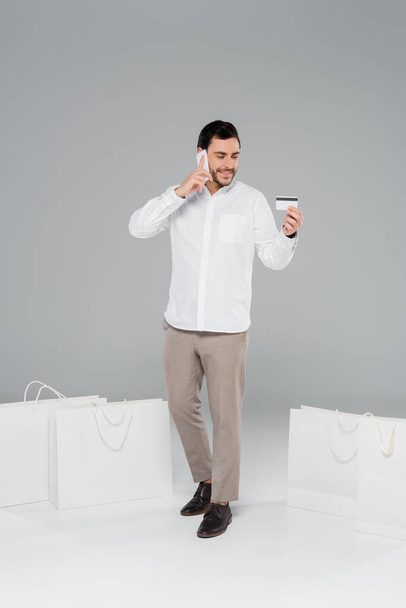 Lächelnder Mann schaut auf Kreditkarte und telefoniert in der Nähe von Einkaufstüten auf grauem Hintergrund - Foto, Bild