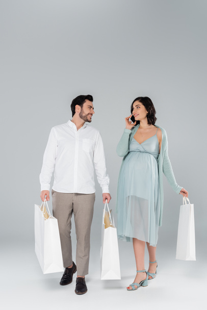 Pregnant woman talking on smartphone near husband with shopping bags on grey background - Fotoğraf, Görsel