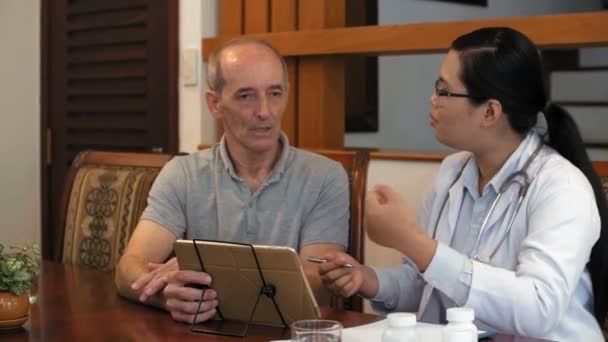 Seurantaväline laukaus naispuolisesta aasialaisesta lääkäristä laboratoriotakissa ja laseissa tabletin tietokoneella ja esittämällä kysymyksiä kypsälle miespotilaalle - Materiaali, video