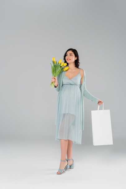 Ευτυχισμένη έγκυος γυναίκα κρατώντας τουλίπες και τσάντα ψώνια σε γκρι φόντο - Φωτογραφία, εικόνα