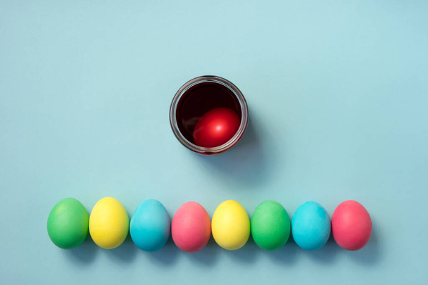 Fila de huevos de Pascua de colores y una taza llena de tinte de huevo sobre fondo azul pastel. Puesta plana. - Foto, Imagen