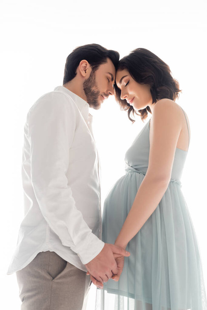 Vista de ángulo bajo de la mujer embarazada con los ojos cerrados cogidos de la mano del marido aislado en blanco - Foto, imagen
