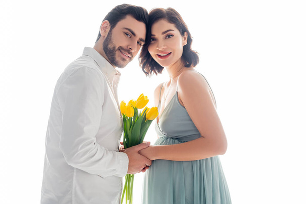 Schwangere Frau mit Blumen lächelt in die Kamera neben Ehemann isoliert auf weiß - Foto, Bild