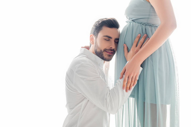 Hombre con camisa escuchando vientre de la esposa embarazada en vestido aislado en blanco - Foto, imagen