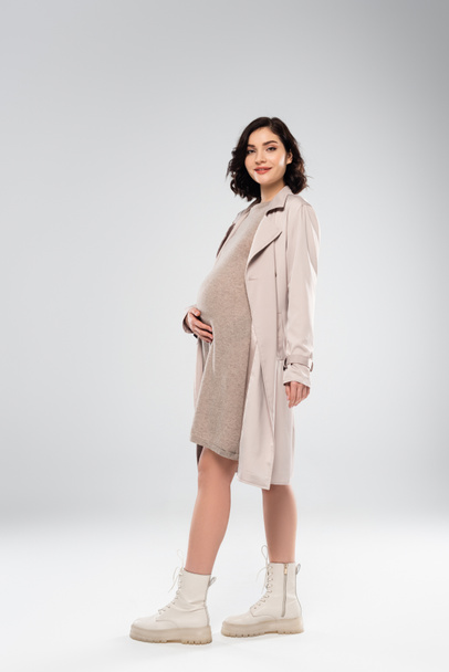Stilvolle schwangere Frau in Mantel und Stiefeln lächelt auf grauem Hintergrund - Foto, Bild