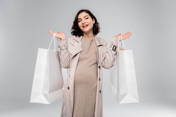 Mujer embarazada con estilo en abrigo sosteniendo bolsas de compras y sonriendo a la cámara aislada en gris  - Foto, Imagen