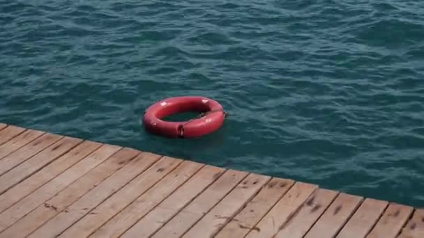 Vörös mentőöv vagy biztonsági gyűrű lebeg a dokk közelében a kék tengervízben. Használatban lévő életmentő felszerelés. Légy óvatos, amikor a mély óceánban úszol, először a biztonság. Mentőmellény, hogy dobja ki a csónakból, ha fulladás - Felvétel, videó