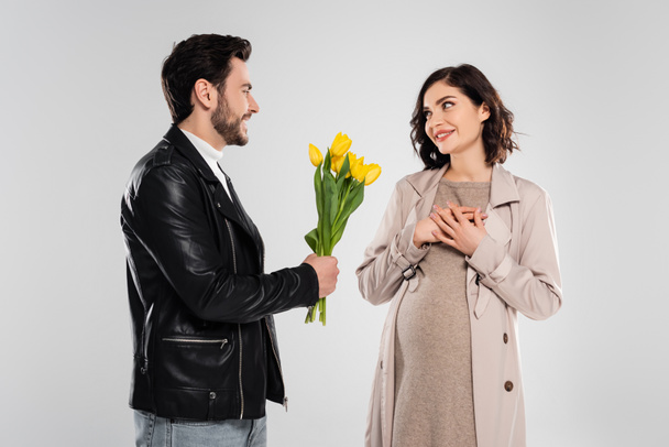Cheerful man holding flowers near pretty pregnant wife on grey background - 写真・画像