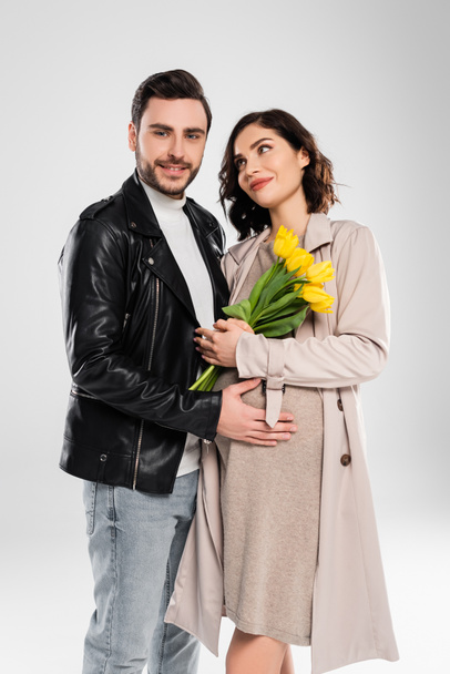 Hombre sonriente en chaqueta de cuero abrazando a la esposa embarazada con tulipanes sobre fondo gris - Foto, imagen