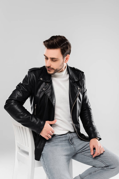 Brunette man in leather jacket sitting on chair isolated on grey - Photo, Image