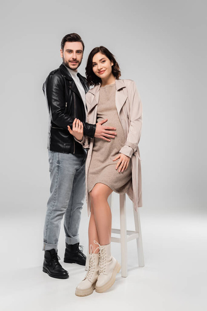 Smiling man touching belly of stylish pregnant wife on chair on grey background  - Foto, immagini