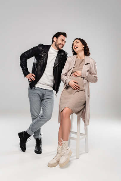 Young man standing near laughing pregnant wife on chair on grey background  - Photo, Image