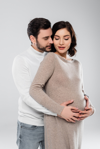 Чоловік обіймає вагітну дружину в одязі ізольовано на сірому
  - Фото, зображення