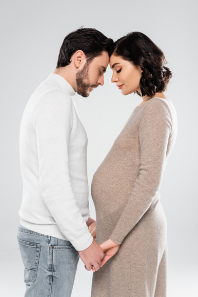 Vista lateral de la mujer embarazada y el marido tomados de la mano aislados en gris  - Foto, imagen