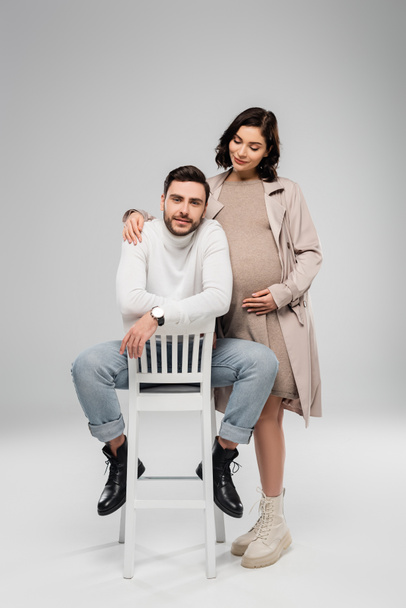 Беременная женщина обнимает мужа, смотрит в камеру на стуле на сером фоне  - Фото, изображение