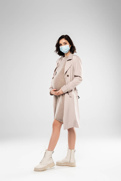 Полная длина стильной беременной женщины в медицинской маске, смотрящей на камеру на сером фоне  - Фото, изображение