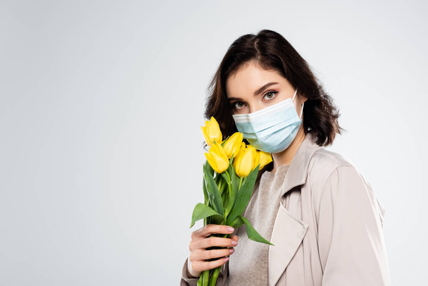 Jeune femme en manteau et masque médical tenant des tulipes isolées sur gris  - Photo, image