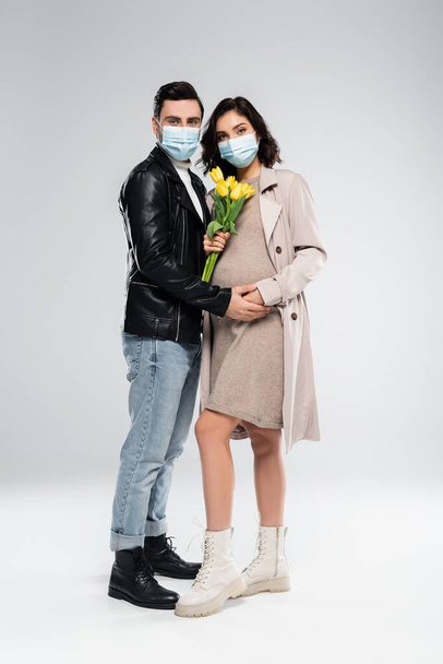 El hombre de estilo en la máscara médica de la mano de la mujer embarazada con flores sobre fondo gris  - Foto, imagen