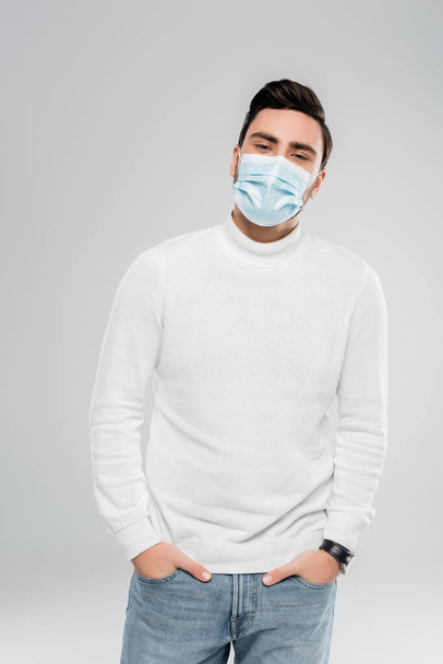 Elegante hombre con máscara médica cogido de la mano en bolsillos de jeans aislados en gris  - Foto, imagen