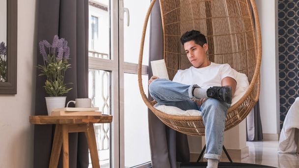 Πορτρέτο του νεαρού άνδρα που διαβάζει ένα βιβλίο, ενώ κάθεται σε μια άνετη καρέκλα στο σπίτι. - Φωτογραφία, εικόνα