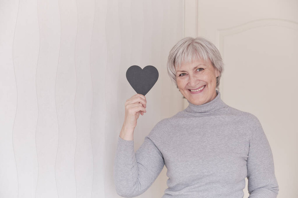 Een portret van een glimlachende oudere blanke vrouwen met kort grijs haar in je hand een klein zwart hart, mockup. - Foto, afbeelding